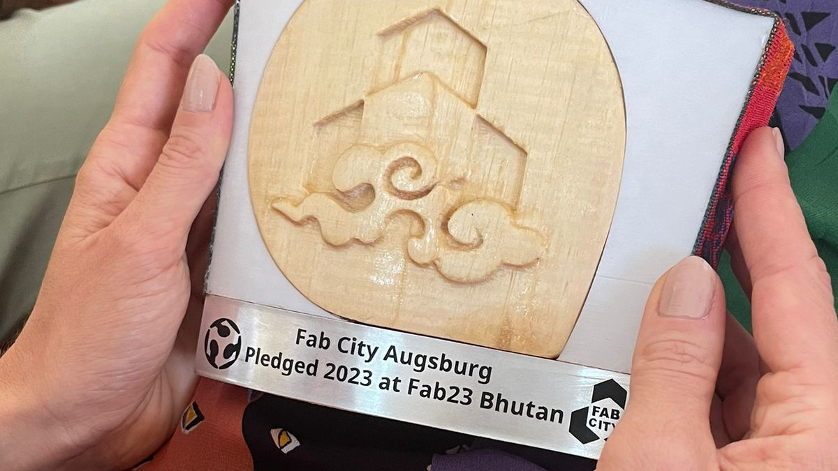 Eine kleine Trophäe aus Holz und Metall für Augsburg, weil sie Deutschlands zweite Fab City wird. Sie wird von zwei Händen gehalten. Auf ihr steht: Fab City Augsburg Pledged 2023 at Fab23 Bhutan.