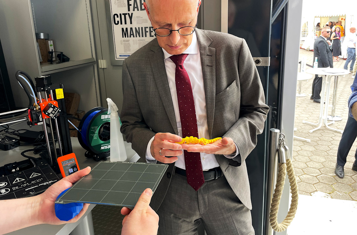 Mayor Peter Tschentscher holds a freshly 3D printed gecko in his hand. The 3D print bed can still be seen and is held by two hands.