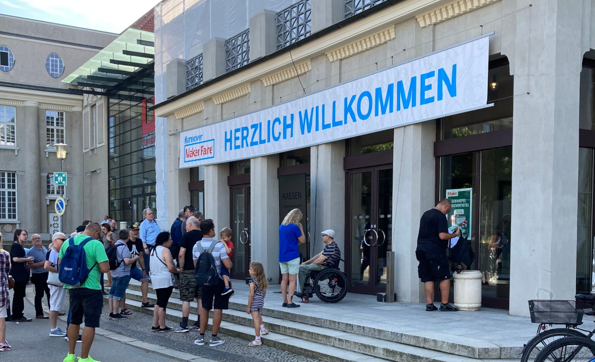 A large gathering of people walking to the entrance of the building where the Maker Faire takes place. Above the entrances hangs a banner with the logo of the Maker Faire, above it says Hannover and large next to it says herzlich willkommen translated to warmly welcome.